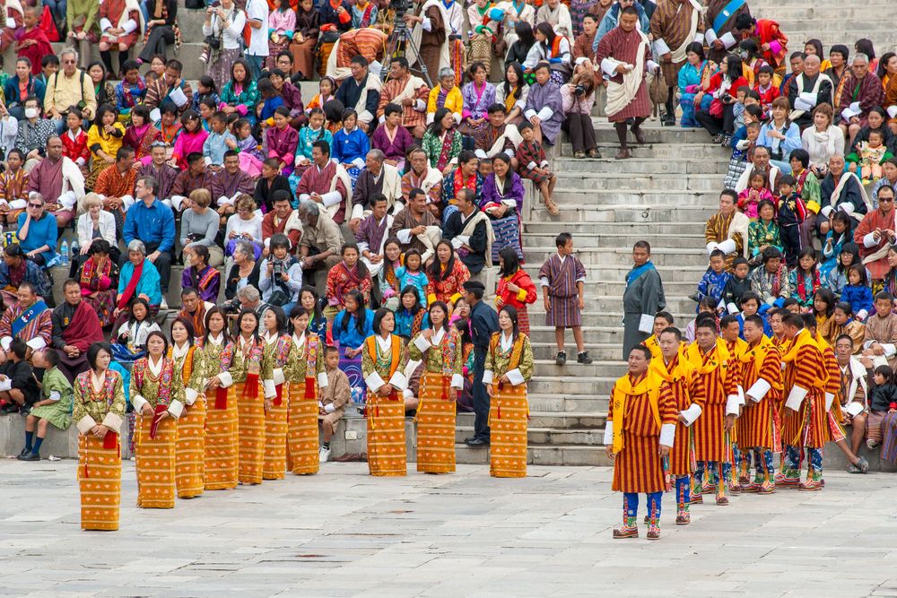 Buddhist monks in traditional orange robes walk in a ceremonial procession past crowds of spectators at a temple.