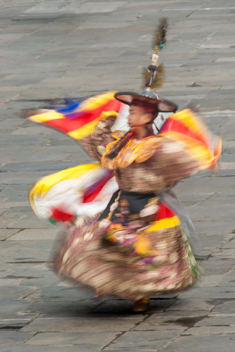 Traditional masked dancer in colorful robes performs a spinning ritual dance with dynamic motion blur on stone ground.