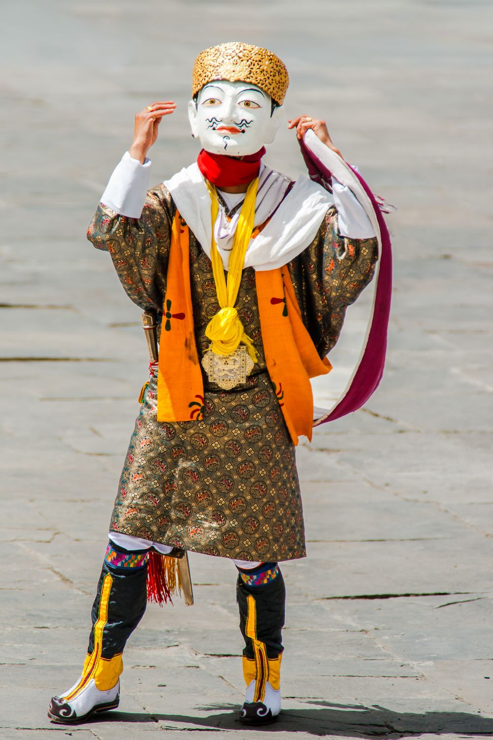 Performer in traditional Bhutanese costume with orange vest and white mask dancing during a festival.