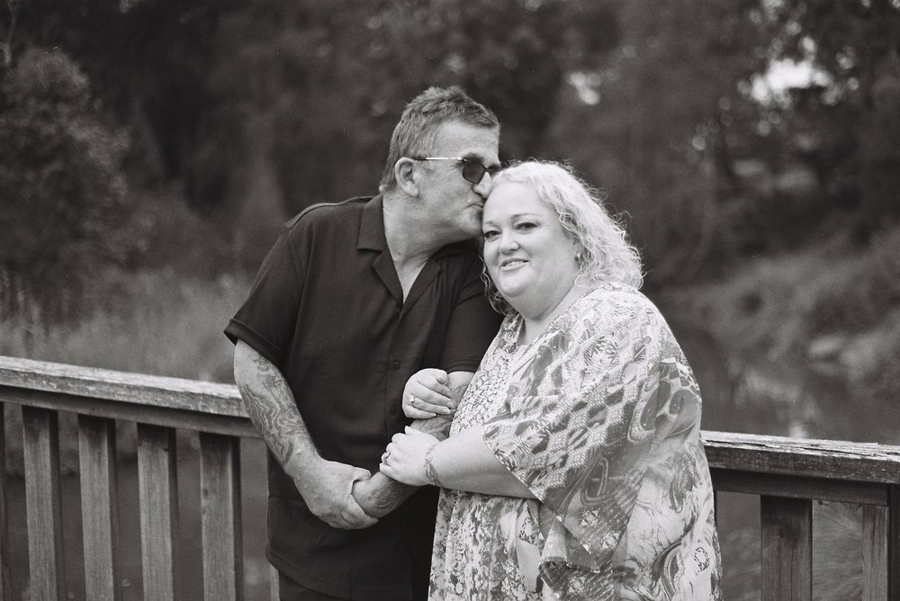 Couple shares tender moments together in black and white photo series on wooden bench in outdoor setting.