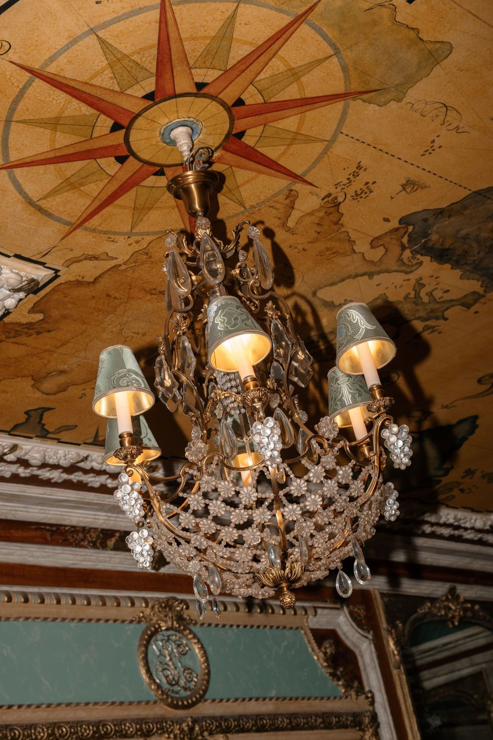 An ornate crystal chandelier hangs from a painted ceiling with classical architectural details.