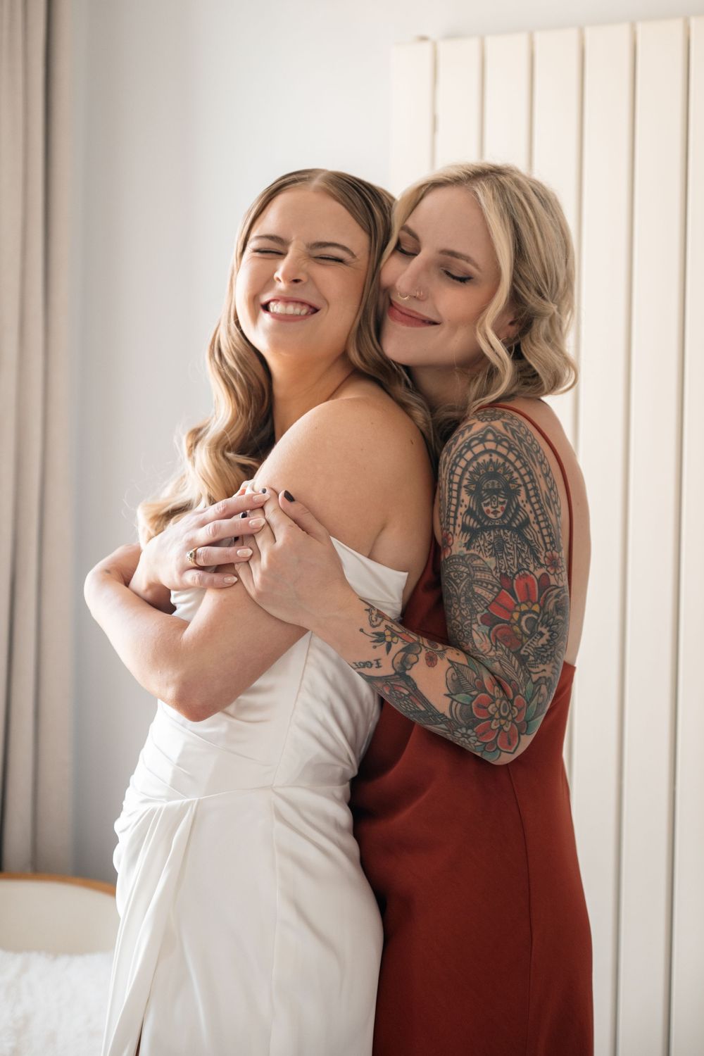 Two women share an intimate embrace in a sun-lit room with white curtains.