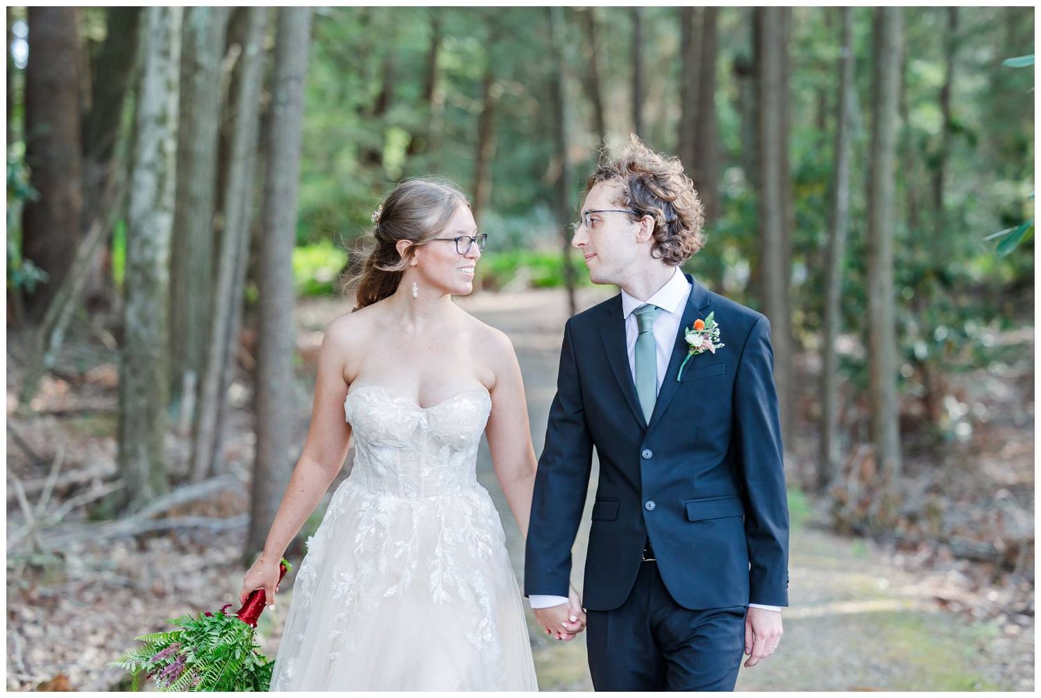 A couple holding hands and smiling at each other while walking in a wooded forest setting.
