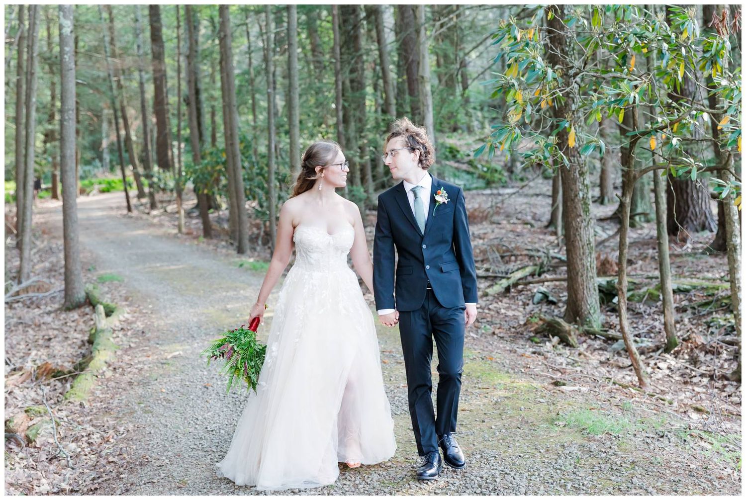 Newlyweds walk hand in hand down a tree-lined forest path in their wedding attire.