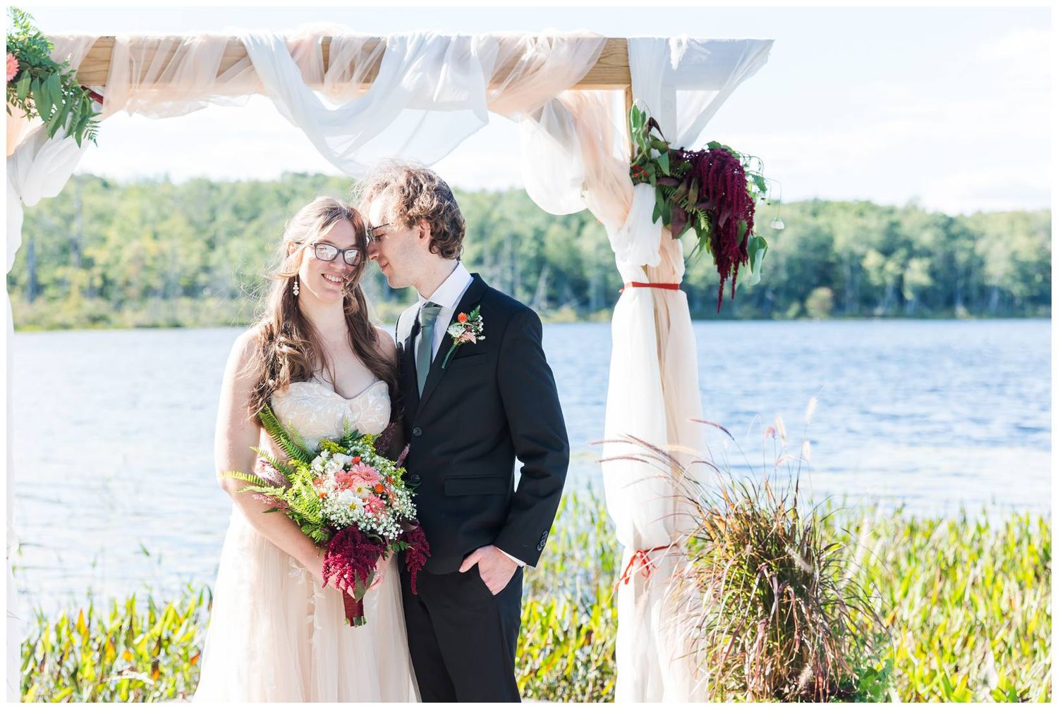 Dreamy waterside wedding portraits with couple standing beside draped white ceremony arch.