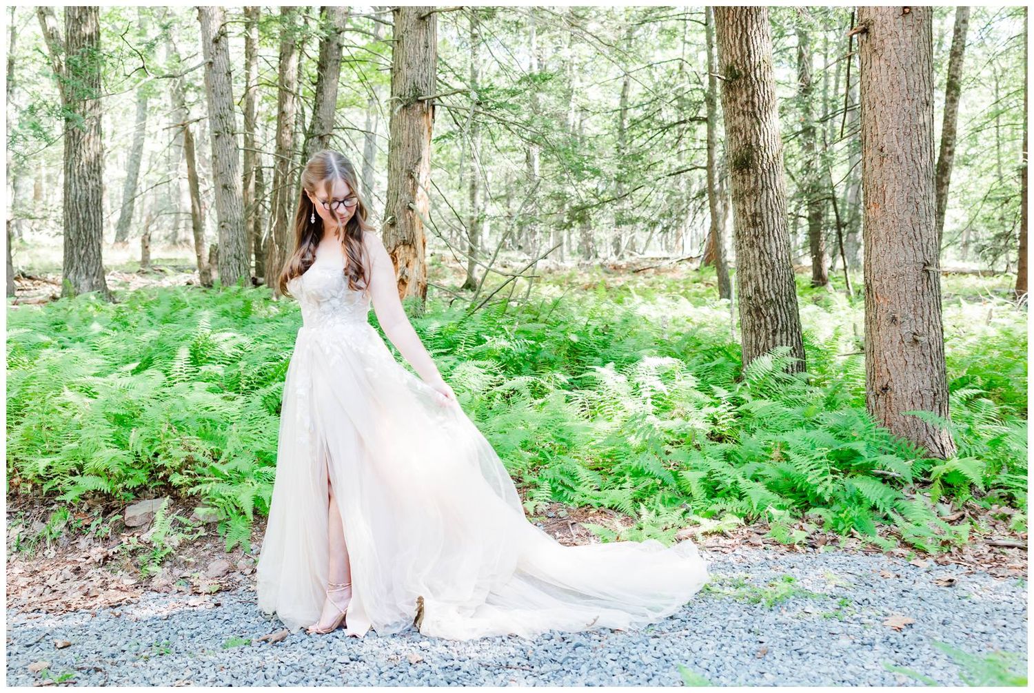 A white wedding dress flows gracefully along a forest path lined with tall pine trees and green foliage.