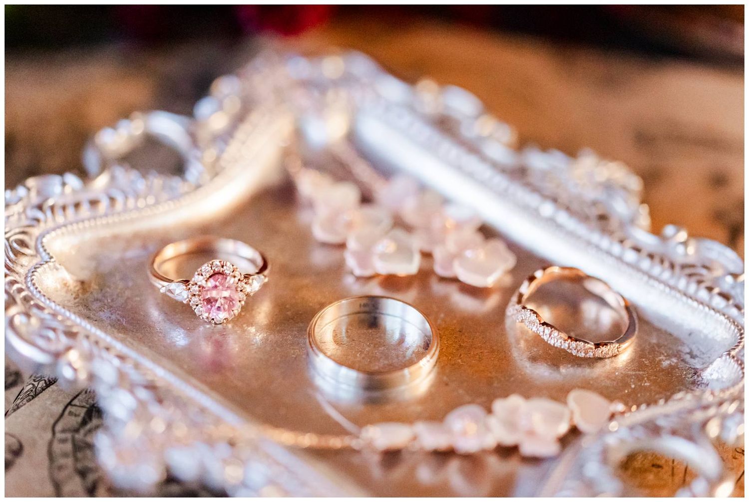Close-up view of gold or silver wedding rings arranged on a wooden surface with pink floral decorations and reflective elements.