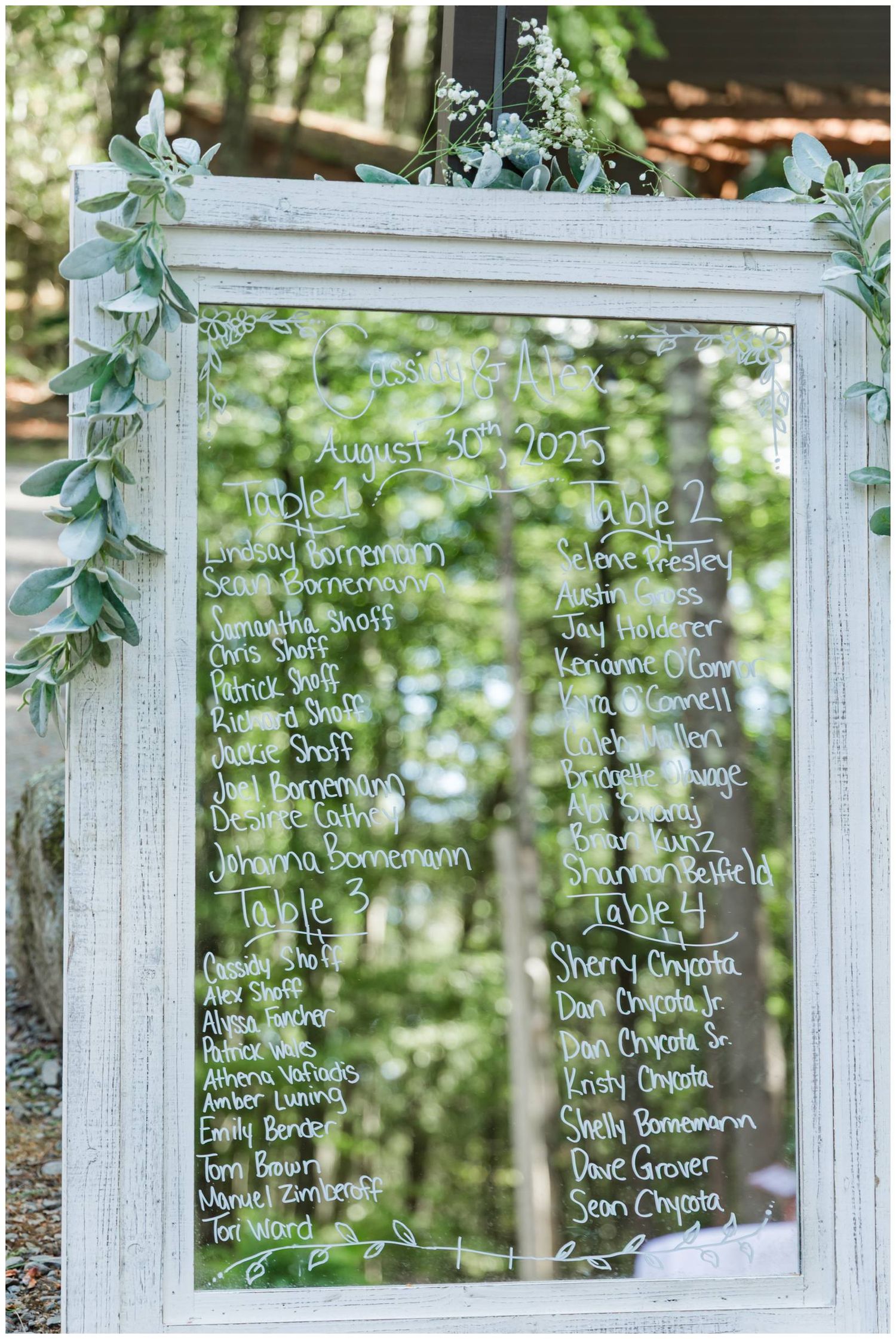 Multiple white-framed mirror seating charts with handwritten guest names and eucalyptus greenery decorations at an outdoor event.