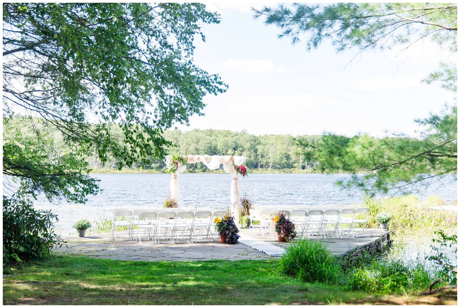 A scenic lakeside wedding ceremony setup with white chairs on the grass, framed by overhanging tree branches.