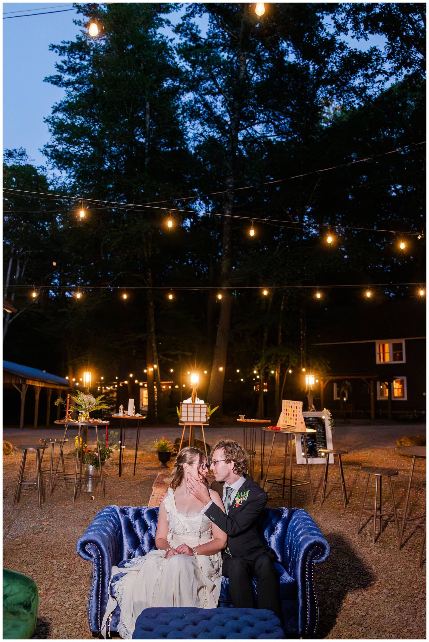 A romantic evening outdoor wedding setting with string lights and a couple seated on a blue velvet sofa.