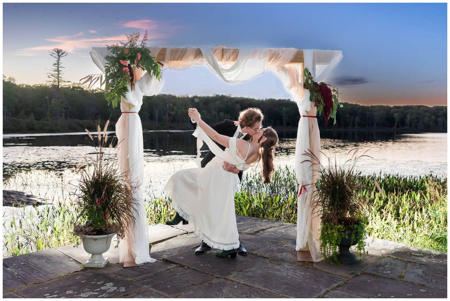 White-clad figures stand by a serene lake at sunset creating a dreamy panoramic wedding scene.