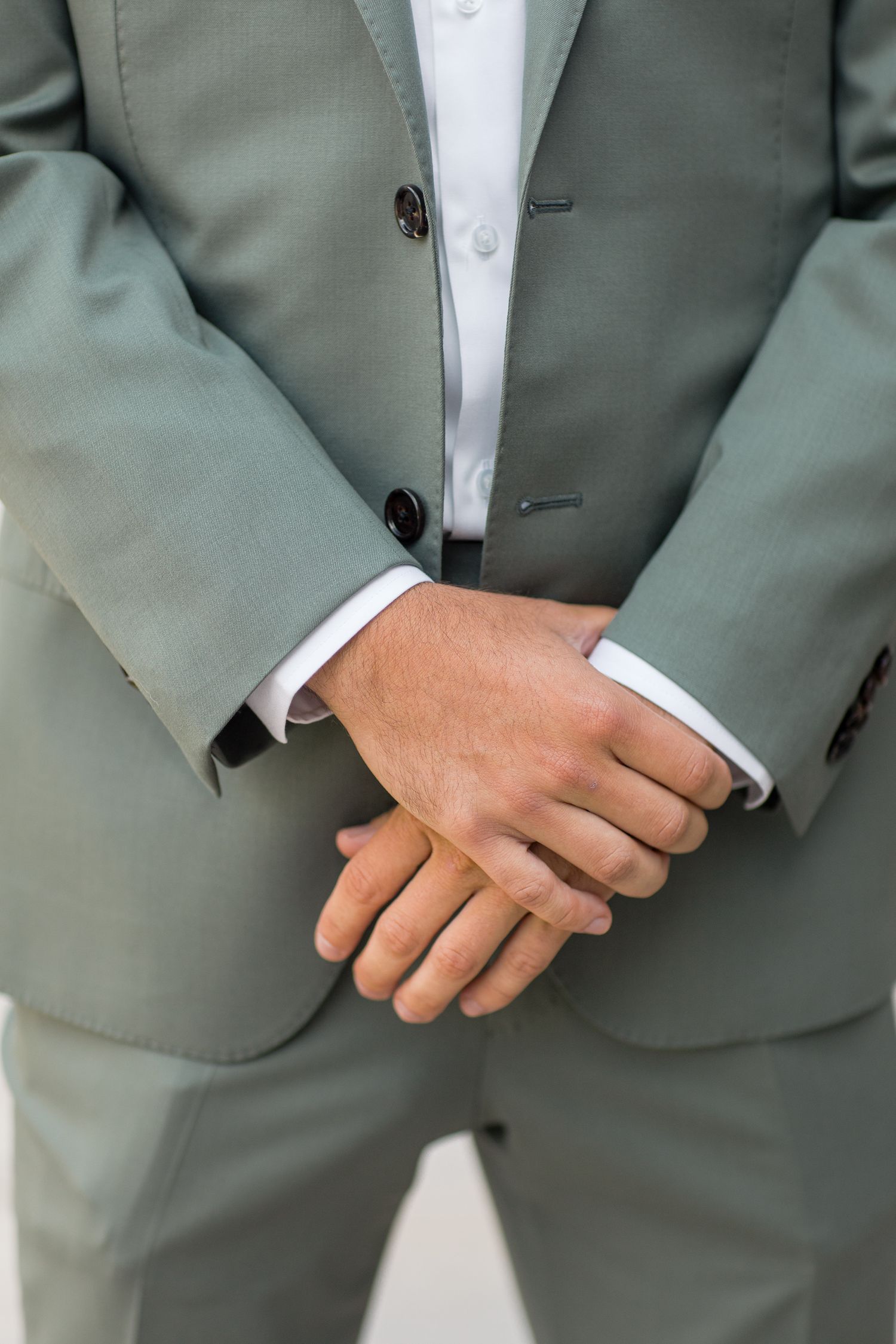 Close-up view of hands clasped over a sage green suit with white dress shirt and black buttons.