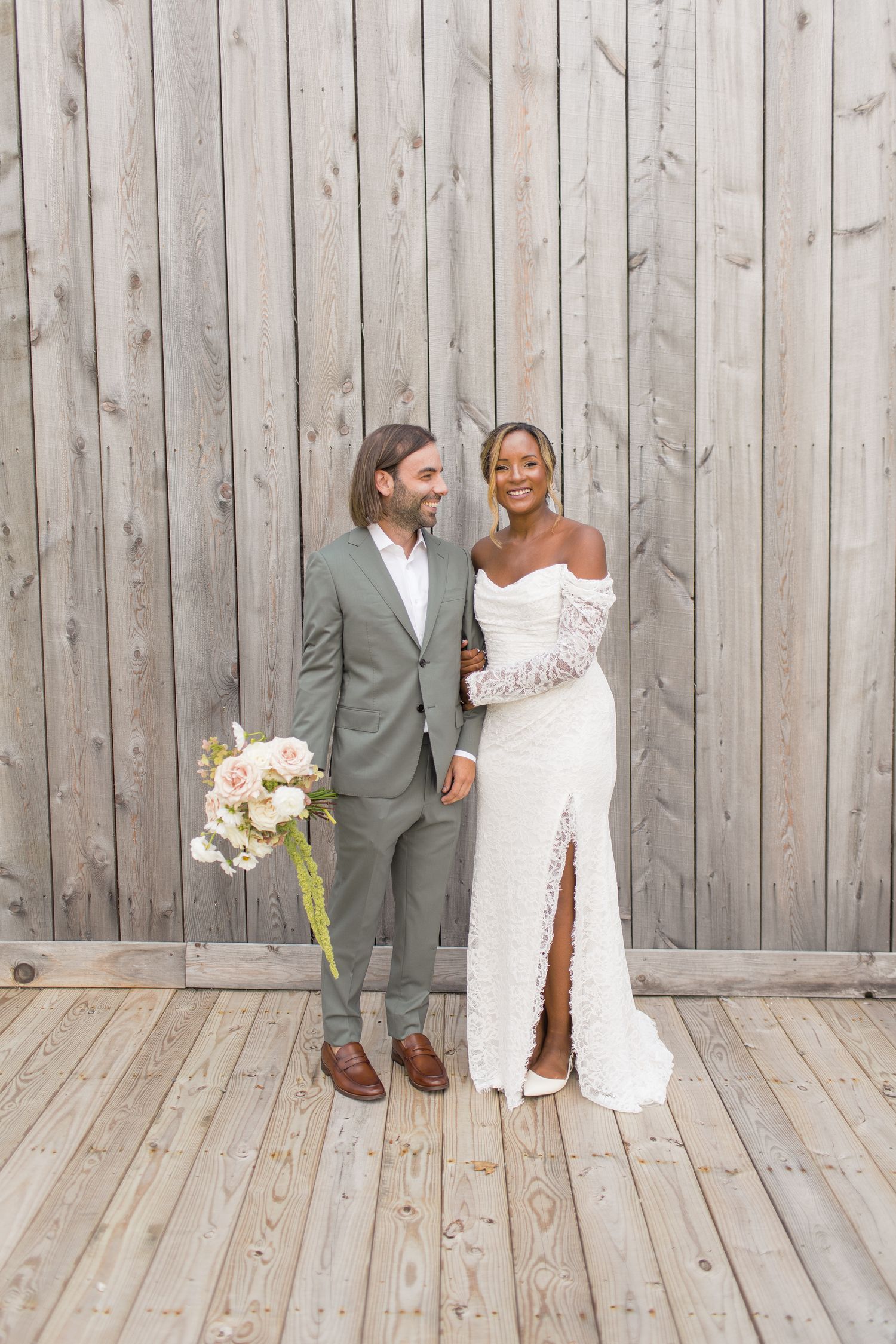A just-married couple in a white lace dress and sage green suit pose together with a pastel floral bouquet against wooden paneling.