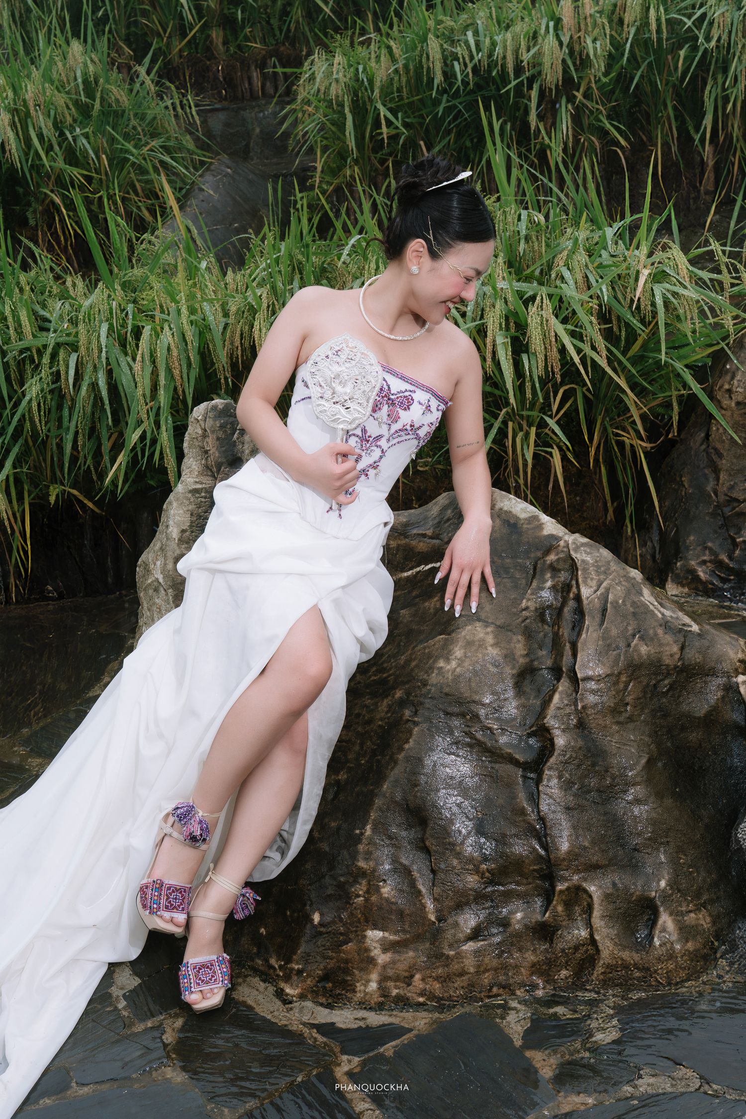 Bride in embroidered strapless gown with slit sits on mossy rocks beside lush green reeds.