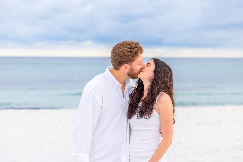 A romantic scene of a couple kissing on a sandy beach with ocean waves and cloudy sky in the background.