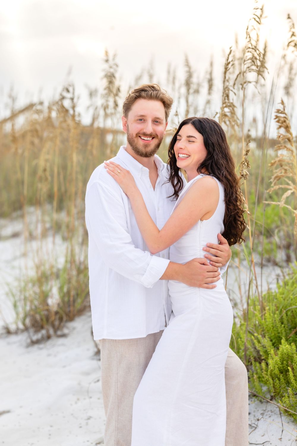 A couple shares intimate moments on a beach with sea oats and sand dunes in the background.