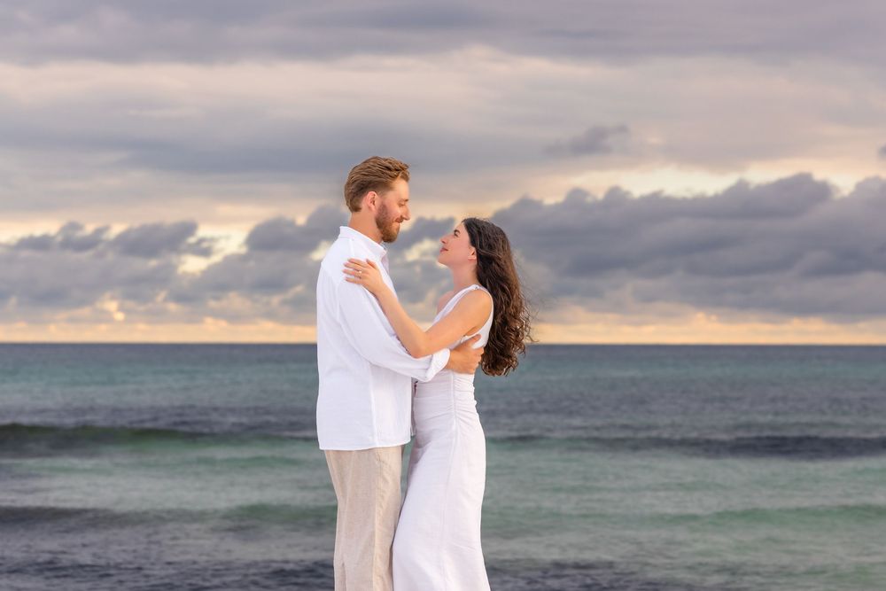 A couple in white outfits dances together on a beach at sunset with waves crashing in the background.