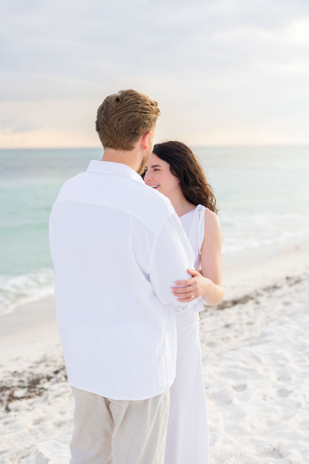 A romantic couple dressed in white embraces on a pristine white sand beach at sunset with turquoise ocean waves nearby.