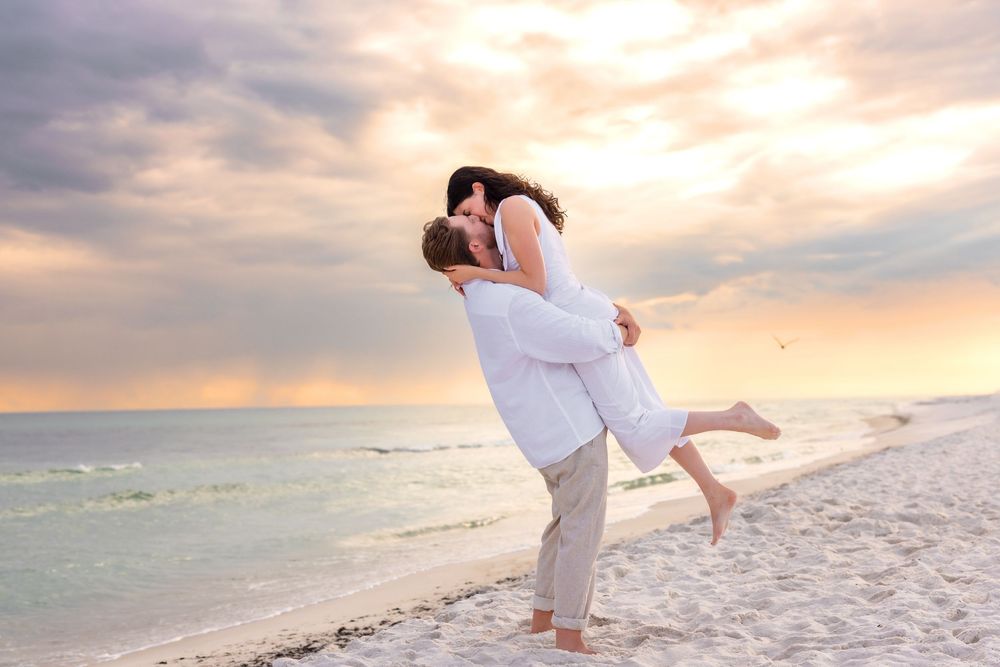 A romantic couple in white attire shares tender moments on a beach during a dreamy sunset photoshoot.