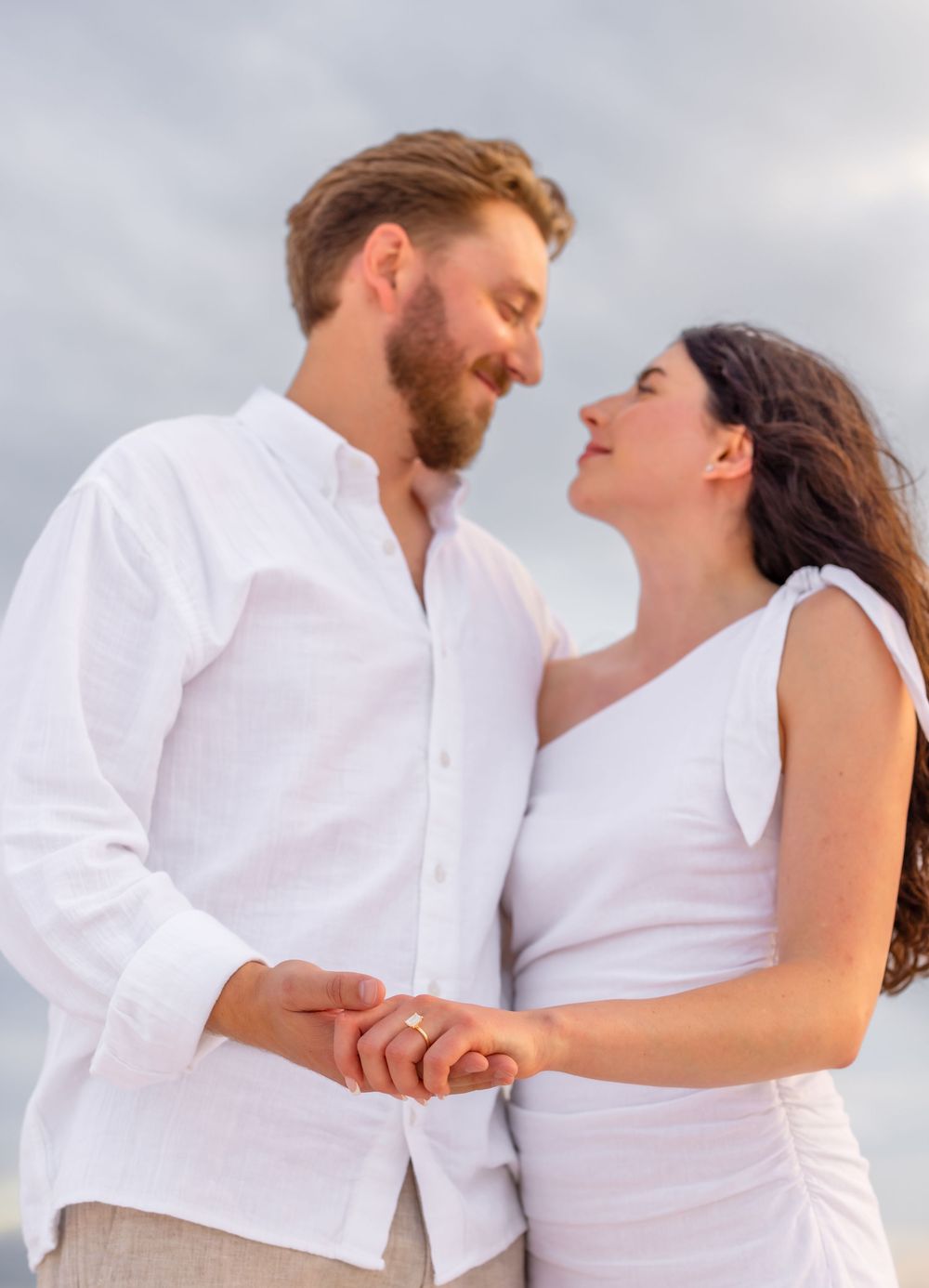 A romantic couple in white clothing share intimate moments during a coastal engagement photo session.