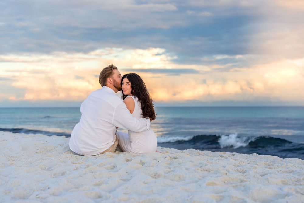 Romantic couple sits together on white sand beach at sunset overlooking the ocean waves.