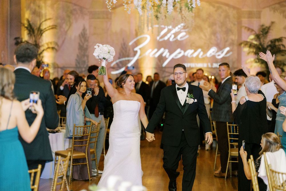 Wedding party dancing together at an elegant reception with chandelier lighting and golden decor.