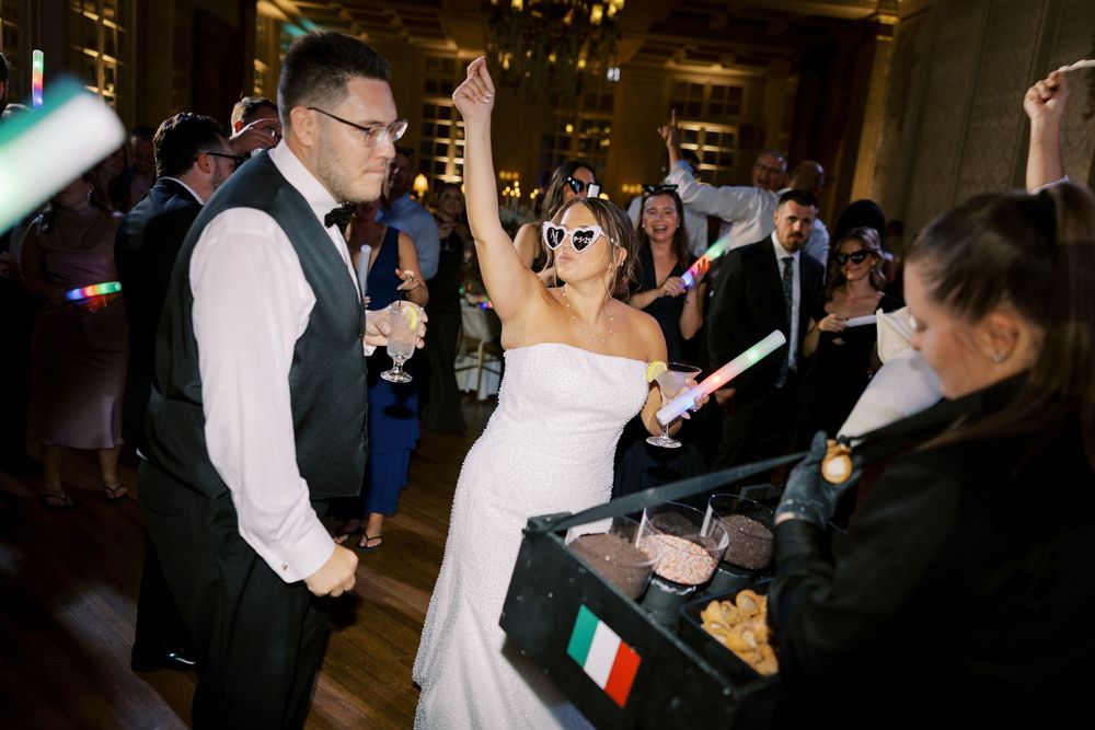 A festive crowd gathers around the newlyweds during an energetic wedding reception celebration with Italian flags.