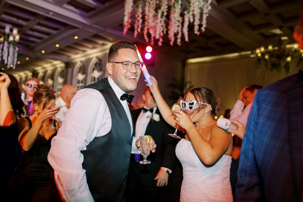 Wedding guests dance enthusiastically on a dimly lit dance floor under sparkling lights at an evening reception.