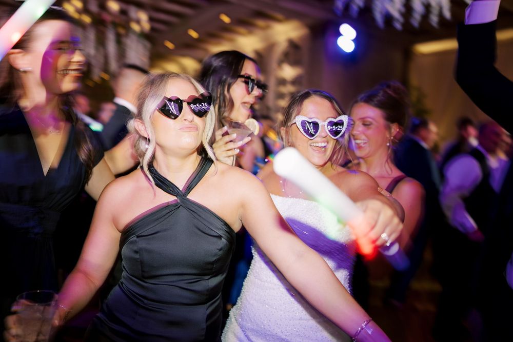 Wedding party guests dance in dark venue wearing heart shaped sunglasses in festive celebration.