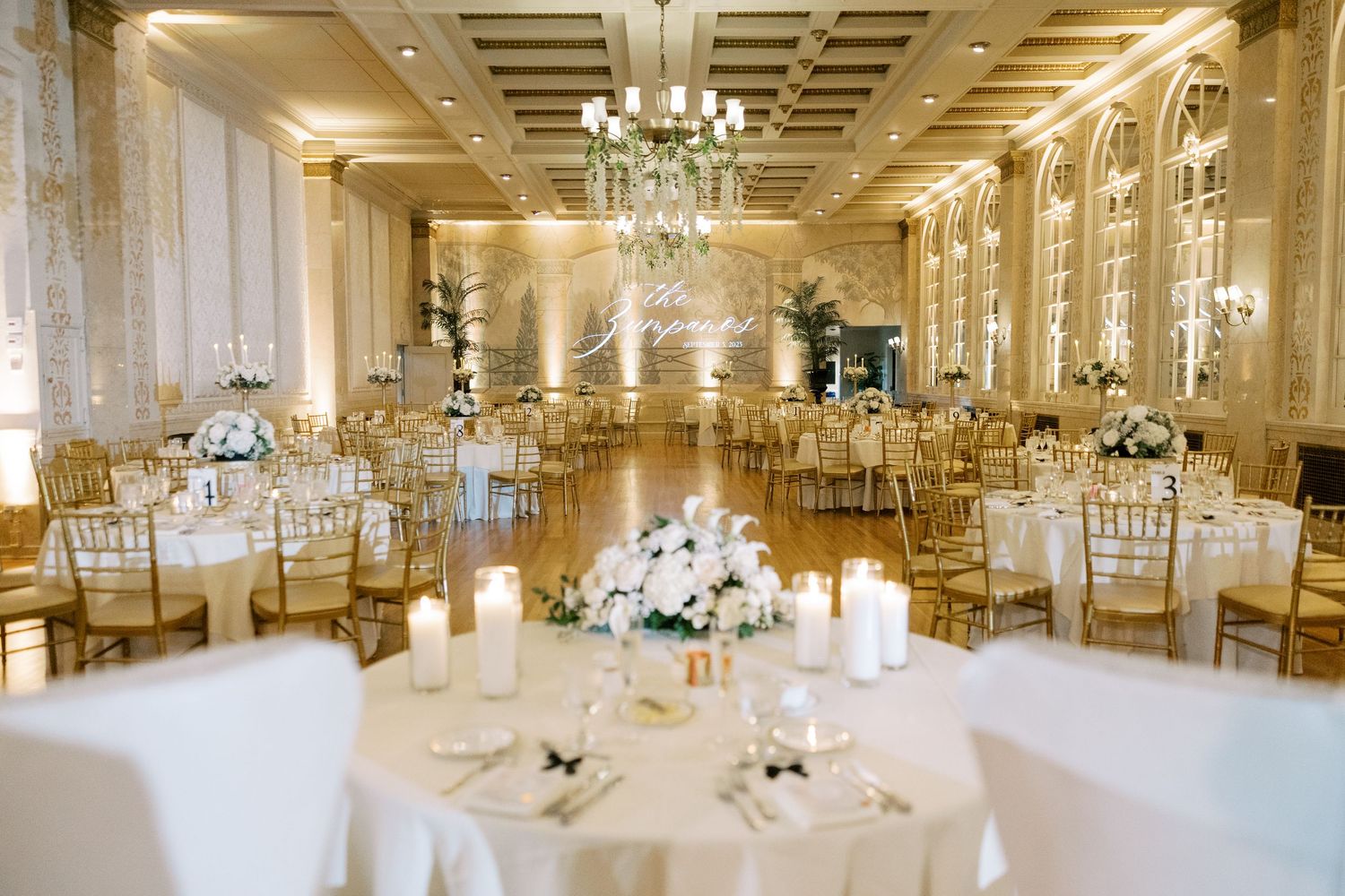Elegant white ballroom venue with crystal chandeliers, draped tables and chairs set up for a wedding reception.