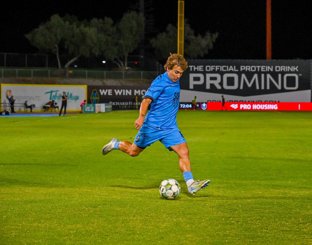 Soccer player in blue kit kicking ball on green field at night with Promino protein drink banner.