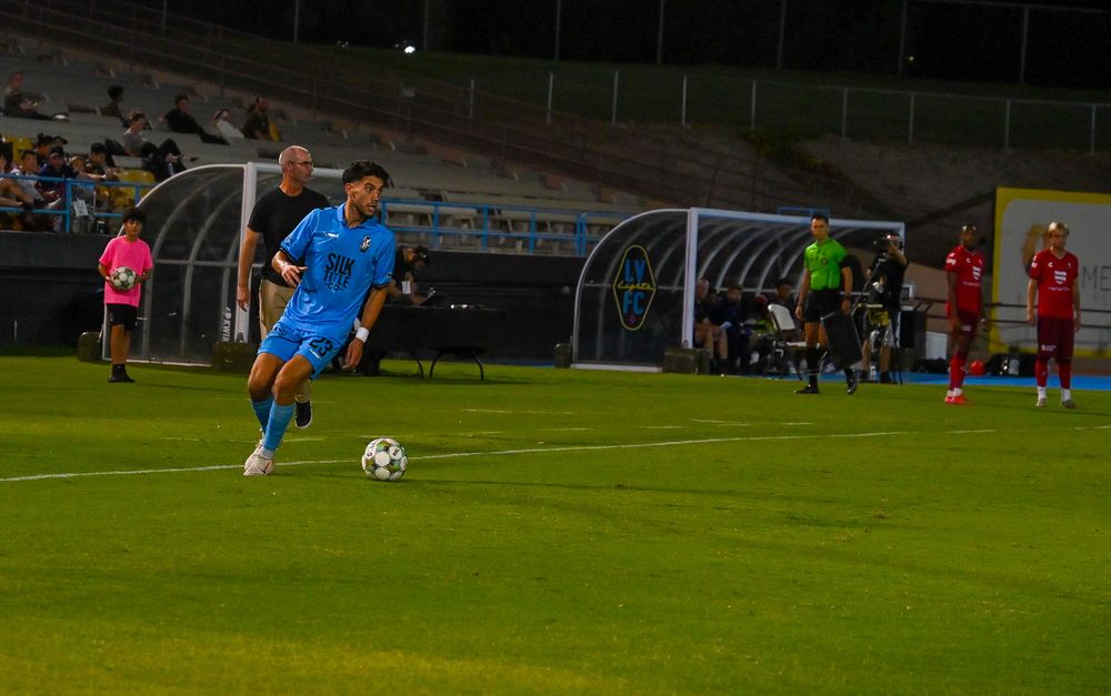 Soccer player in blue uniform dribbles ball near team bench during night match at stadium.