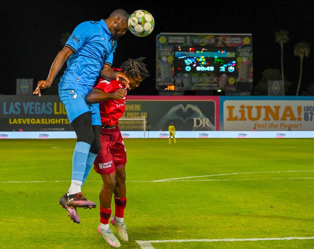 Two soccer players battle for header during night match with Las Vegas Lights scoreboard visible.