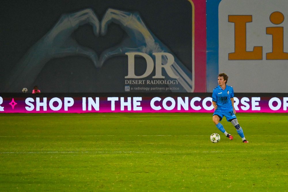 A soccer player in blue uniform controls the ball near Desert Radiologyadvertising boards.
