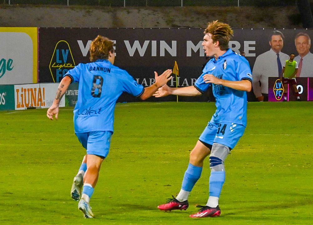Two soccer players in light blue uniforms celebrate goal during night match near sideline banner.