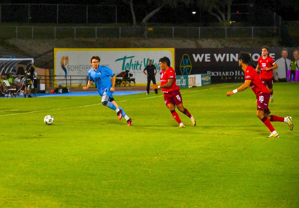 Soccer player in blue sprints past two red-uniformed defenders during night match on green pitch.