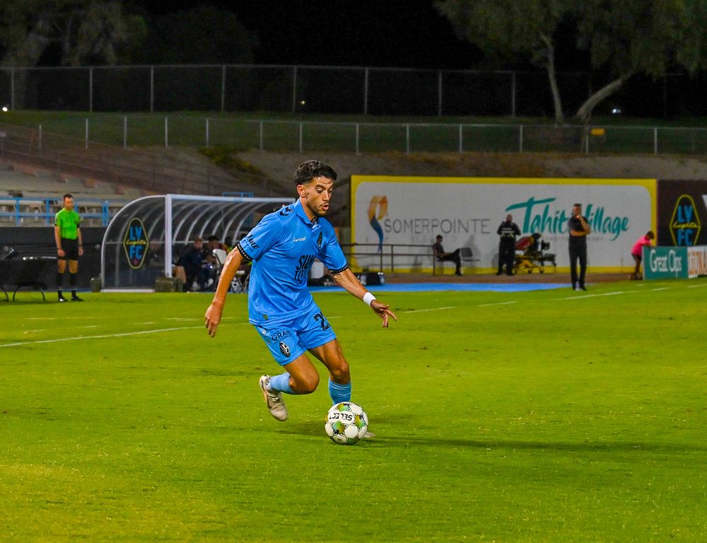 Soccer player in blue uniform dribbling ball during night match with Tahiti Village advertisement.