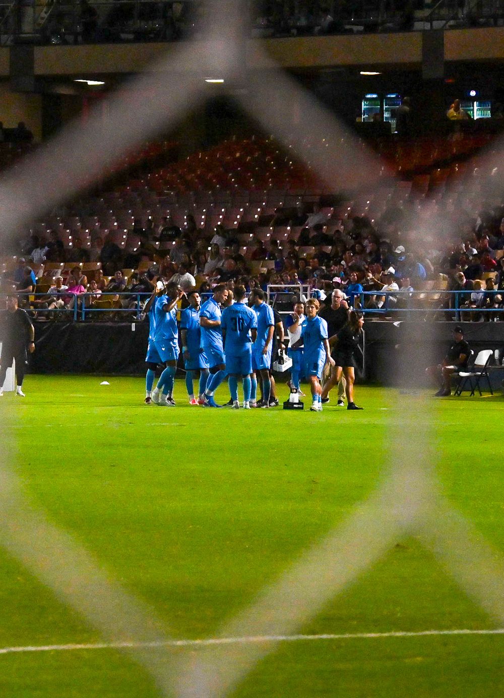 Soccer team in blue uniforms huddles on field during night game viewed through goal netting.