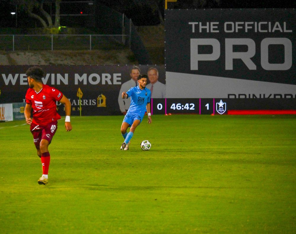 Soccer player in blue dribbles past red-uniformed opponent with 46:42 match clock on scoreboard.