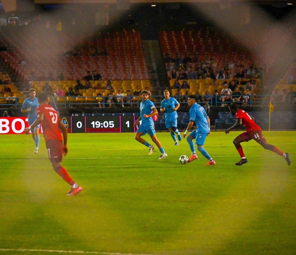 Soccer players in blue and red uniforms compete during night match viewed through goal netting.