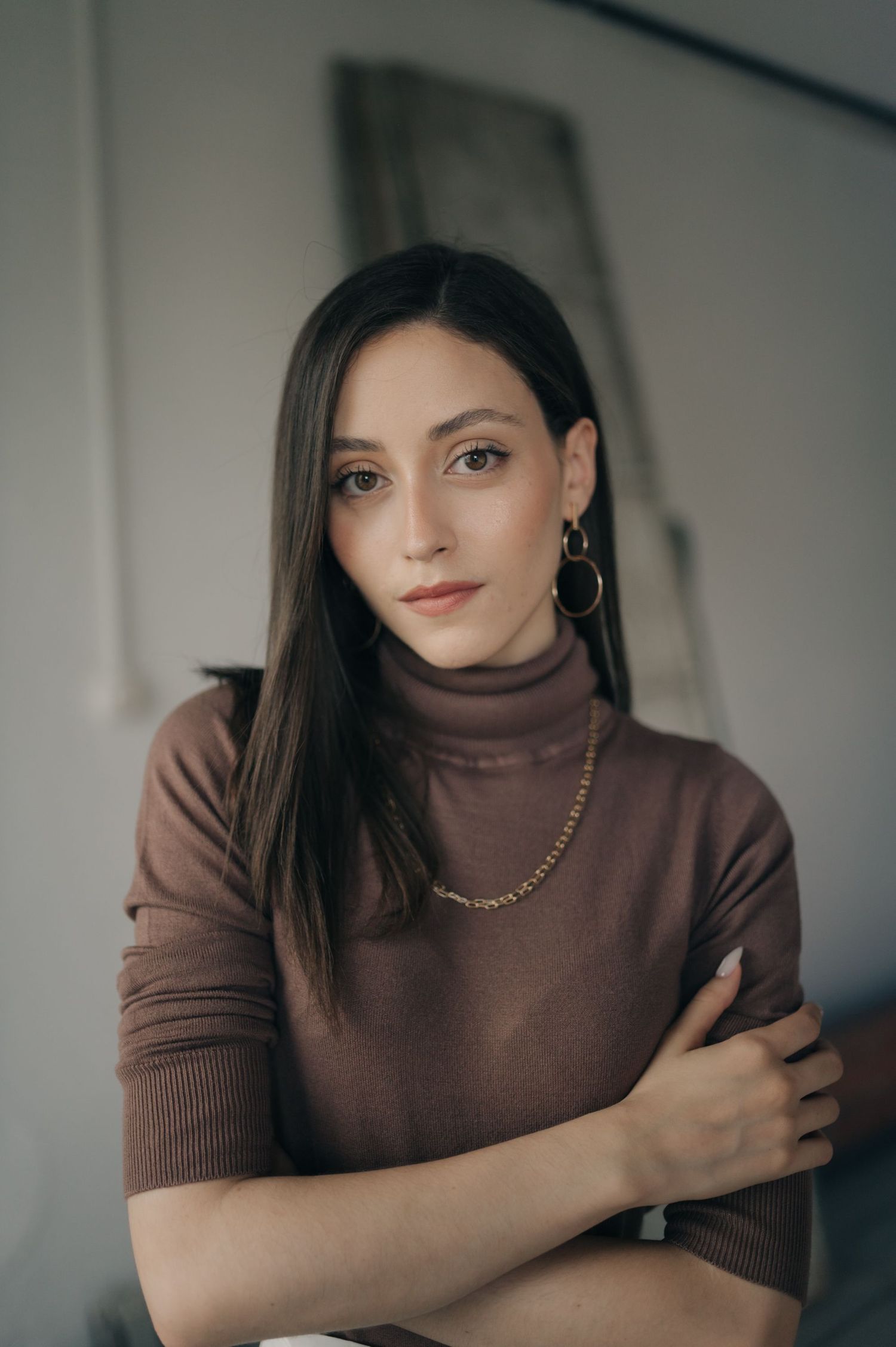 Portrait of person in brown turtleneck sweater with gold jewelry against neutral background.