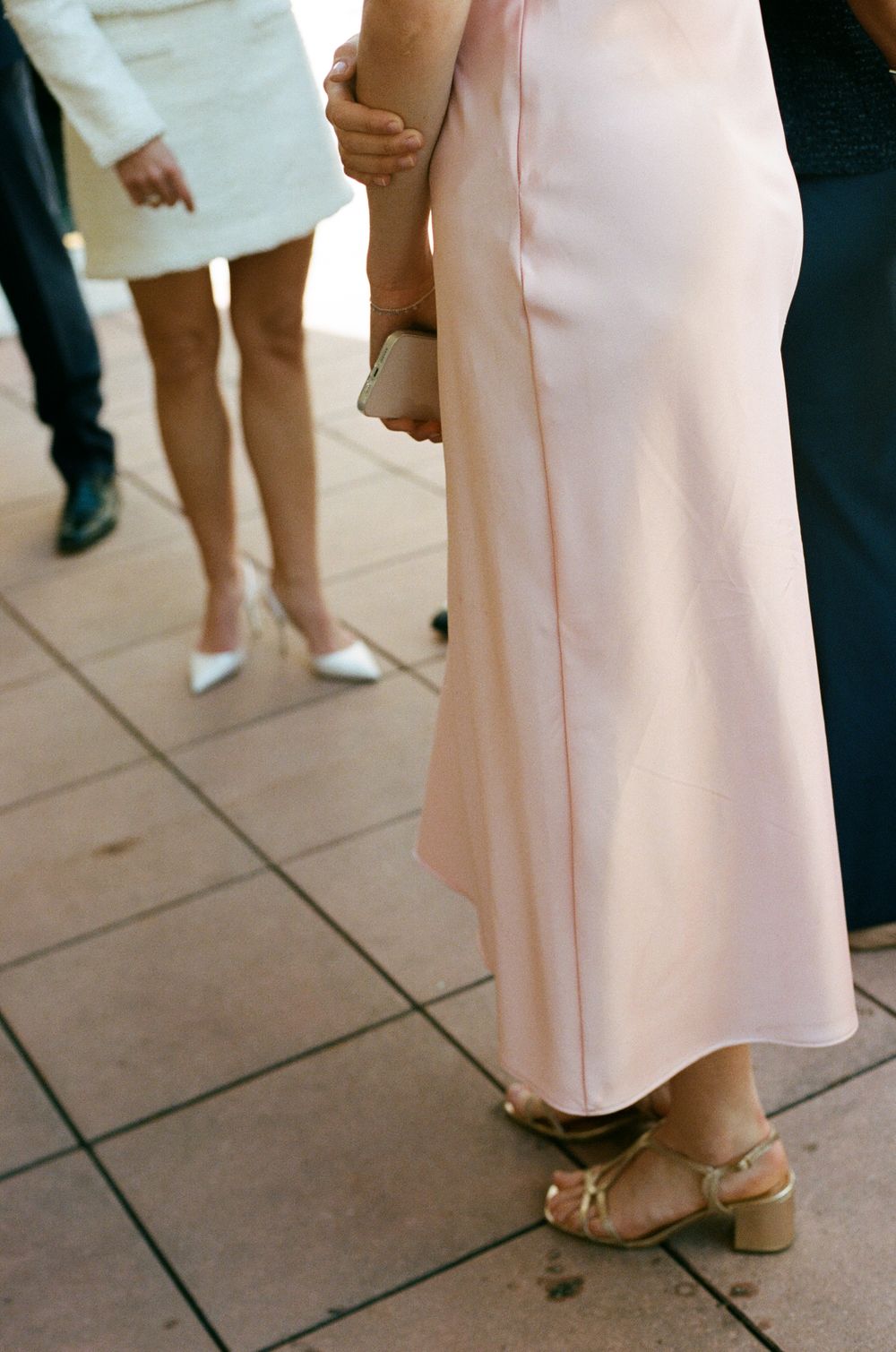 A person wearing a long pink dress and gold sandals stands on textured pavement during a gathering.