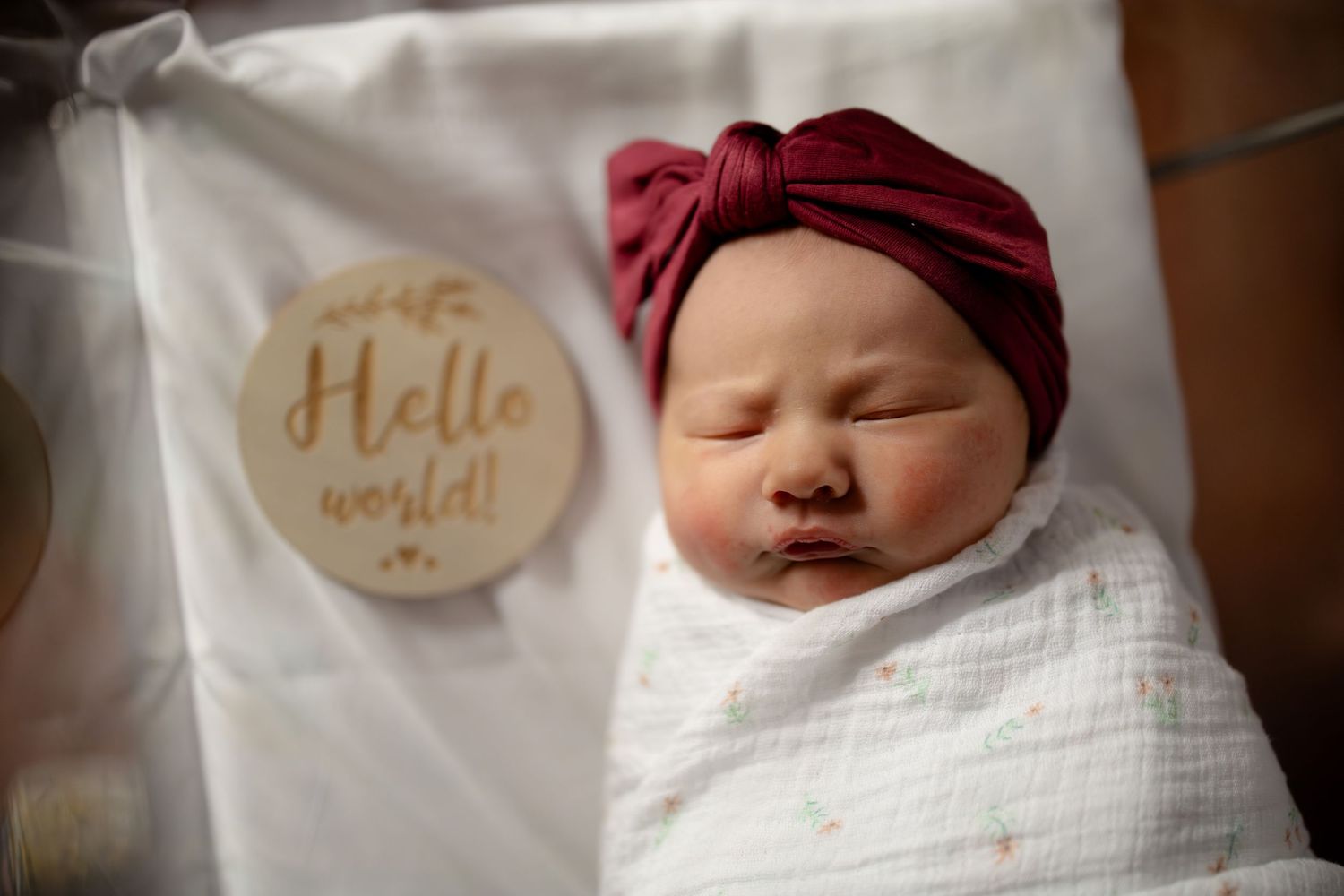 Peaceful newborn baby girl in burgundy bow turban sleeping swaddled beside wooden Hello World milestone disc.