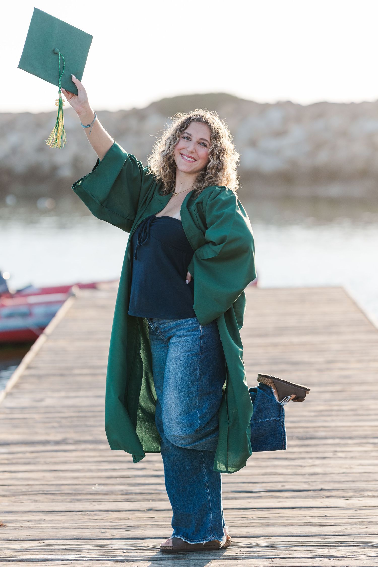 A person in a green coat and jeans poses playfully on a wooden dock during a photoshoot.