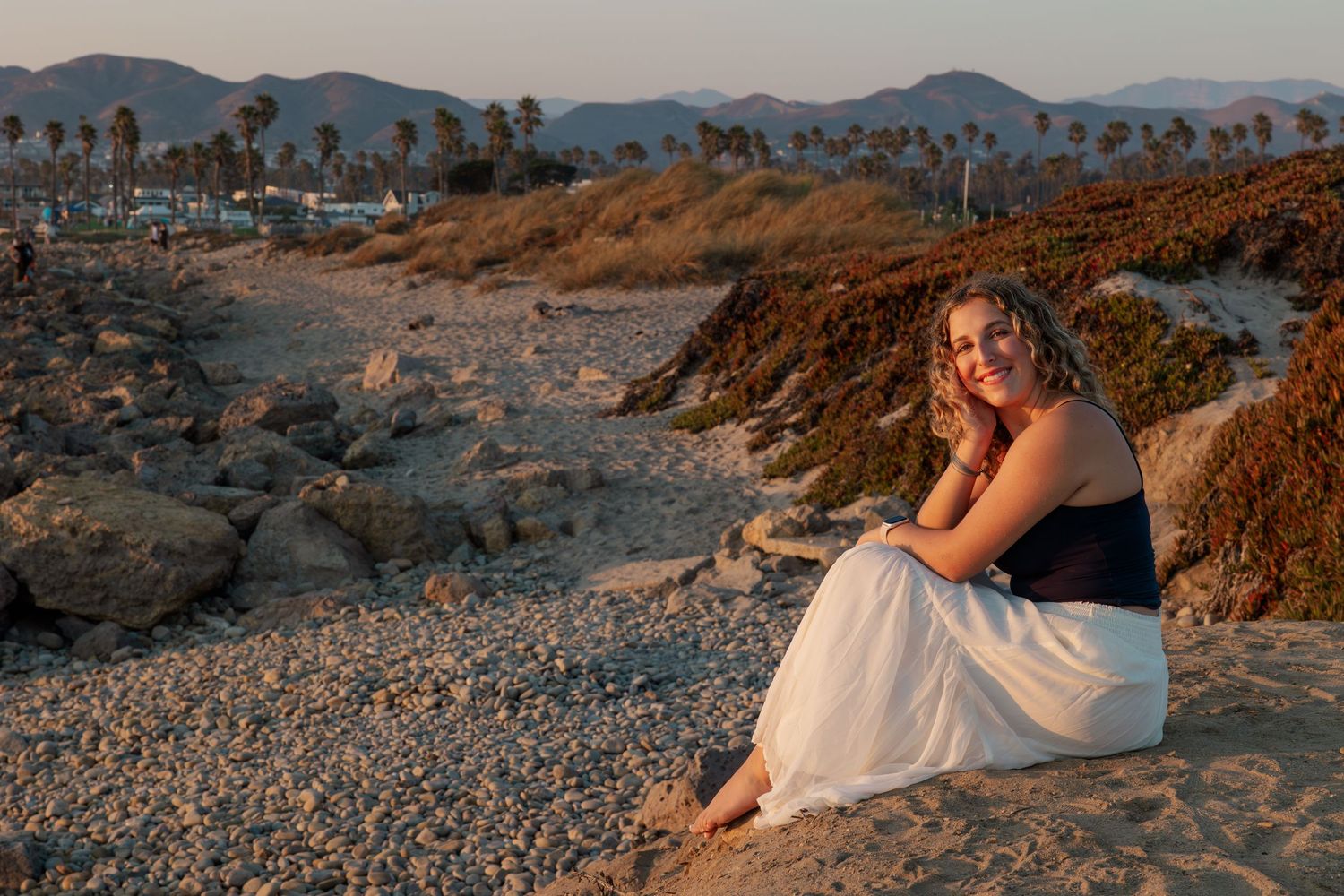 Desert landscape at sunset with mountains in background and person reclining on rocky ground in white dress.