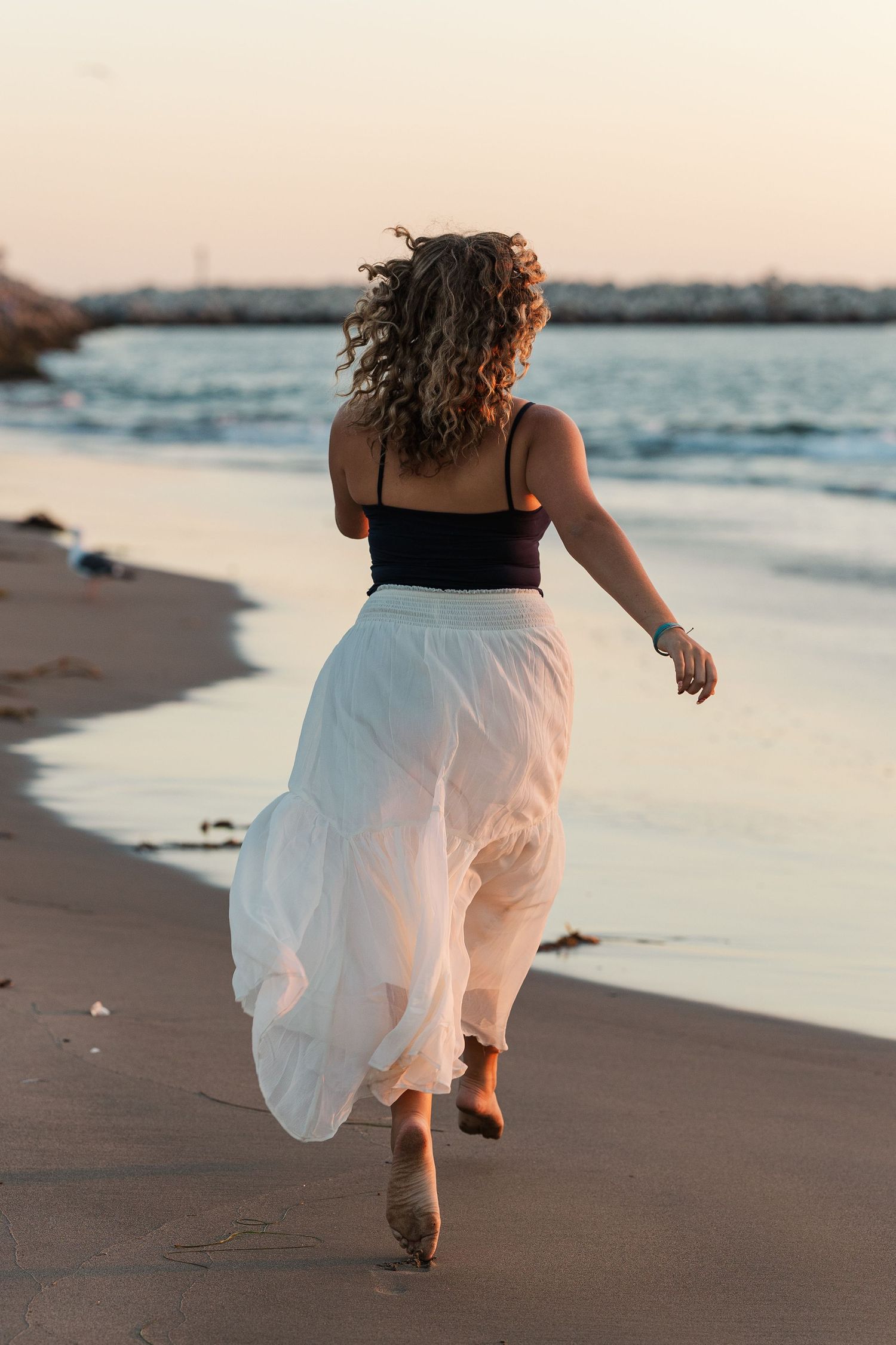Person in a white flowing dress runs barefoot along a sandy beach at sunset with ocean waves in view.