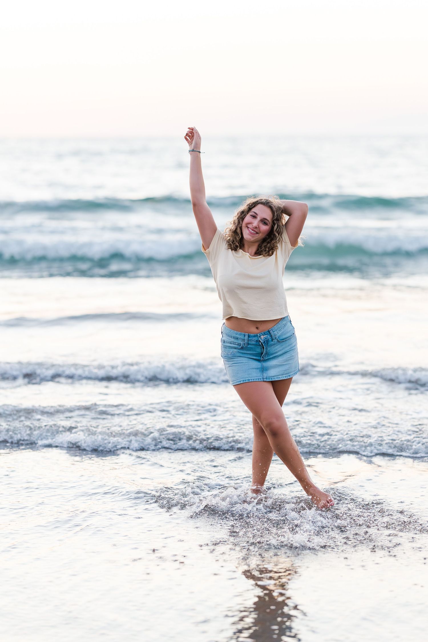 Person splashing and dancing joyfully in the ocean waves at sunset wearing denim shorts.