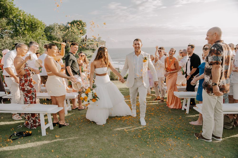 Wedding guests throw confetti and celebrate during an outdoor garden ceremony on a sunny day.
