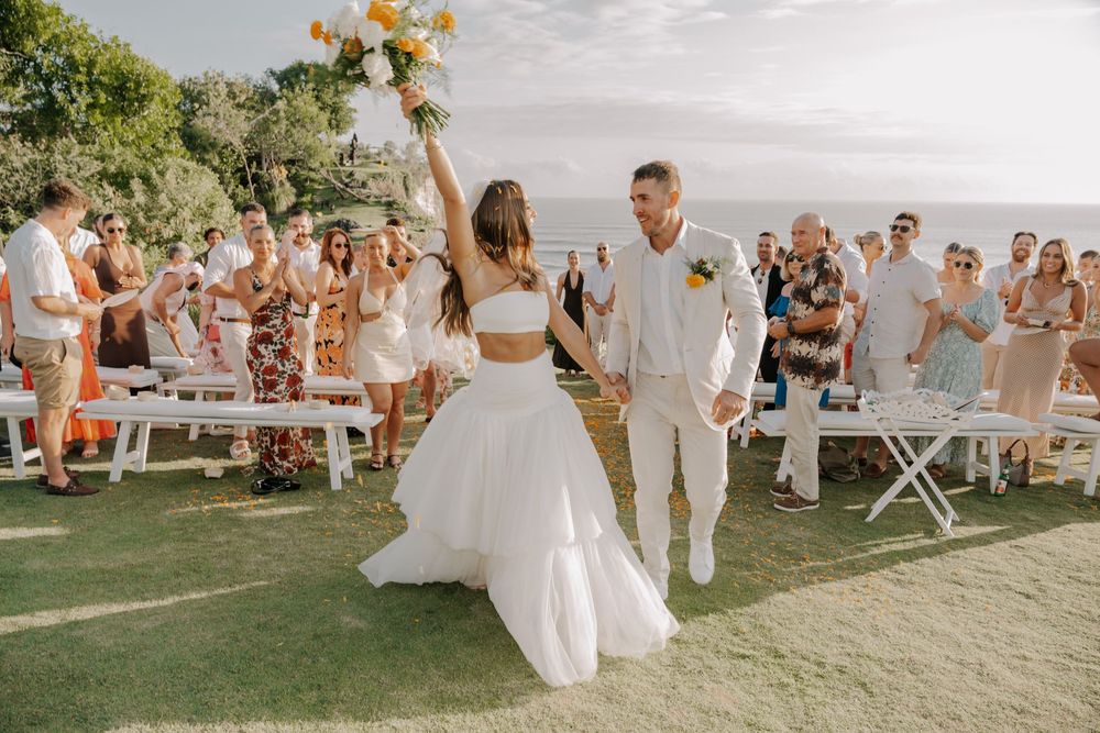 Wedding guests toss flower petals in celebration as the newlywed couple walks down a grassy aisle on a sunny day.