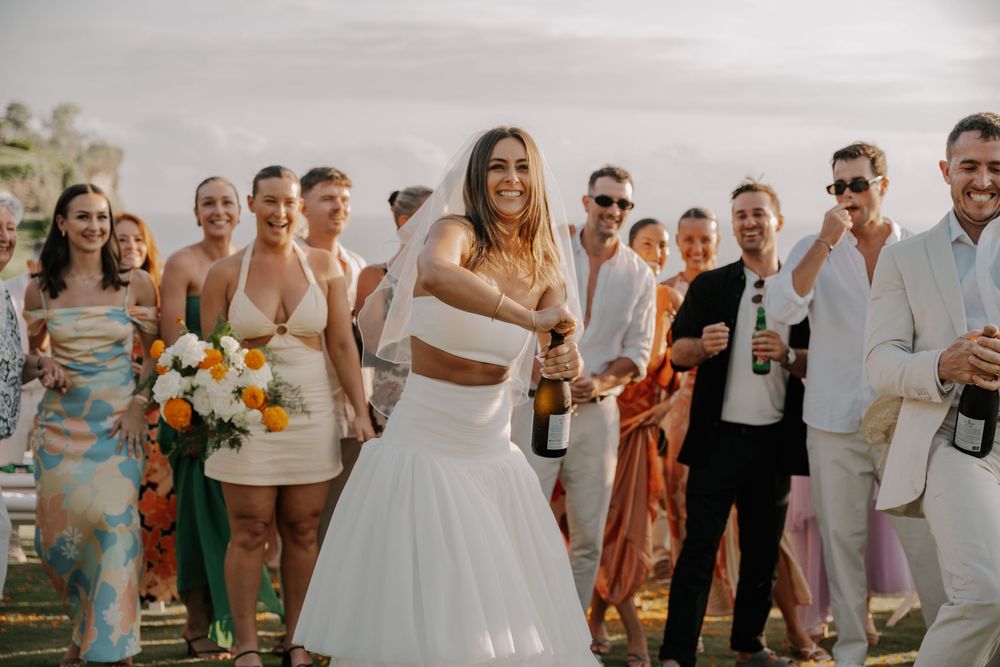 Wedding party members hold drinks and smile while posing together during an outdoor celebration.